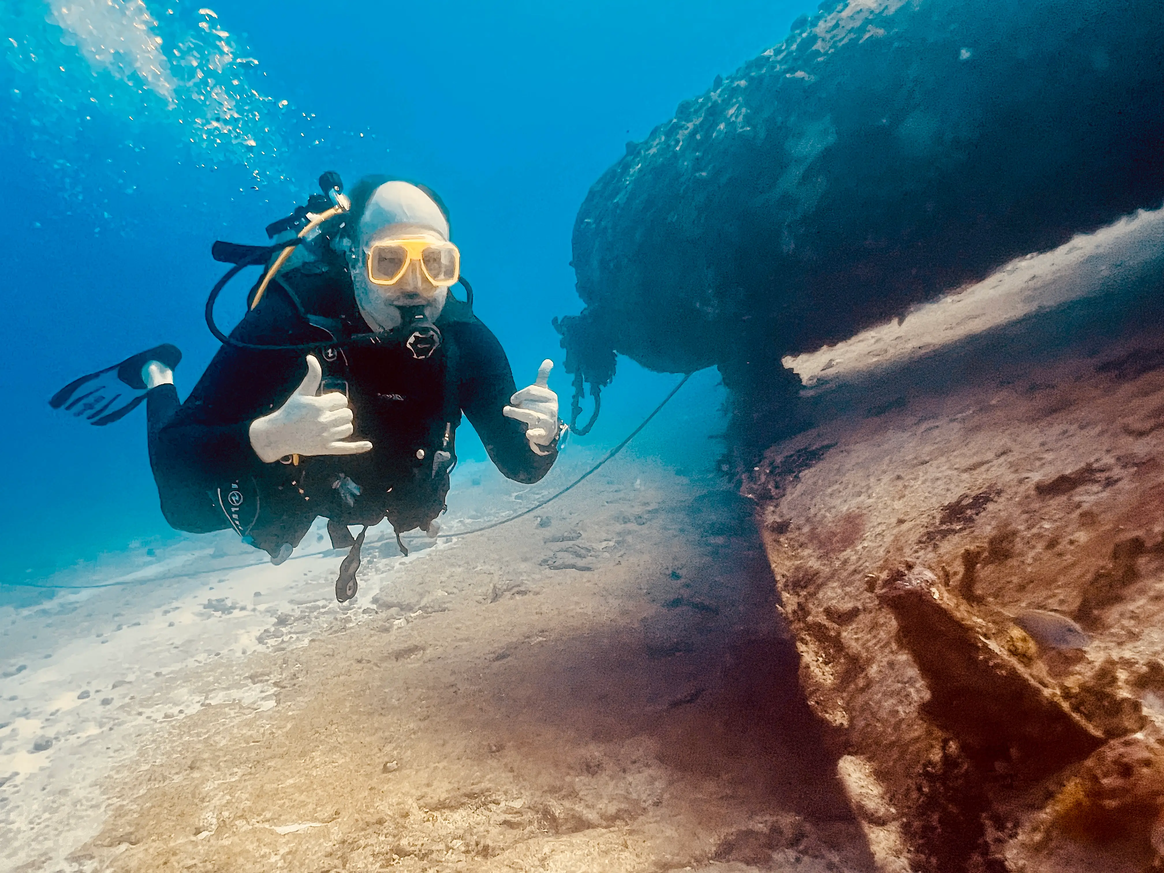 A diver gives a thumbs-up in front of a coral-encrusted wreck, with bubbles rising in clear blue water.
