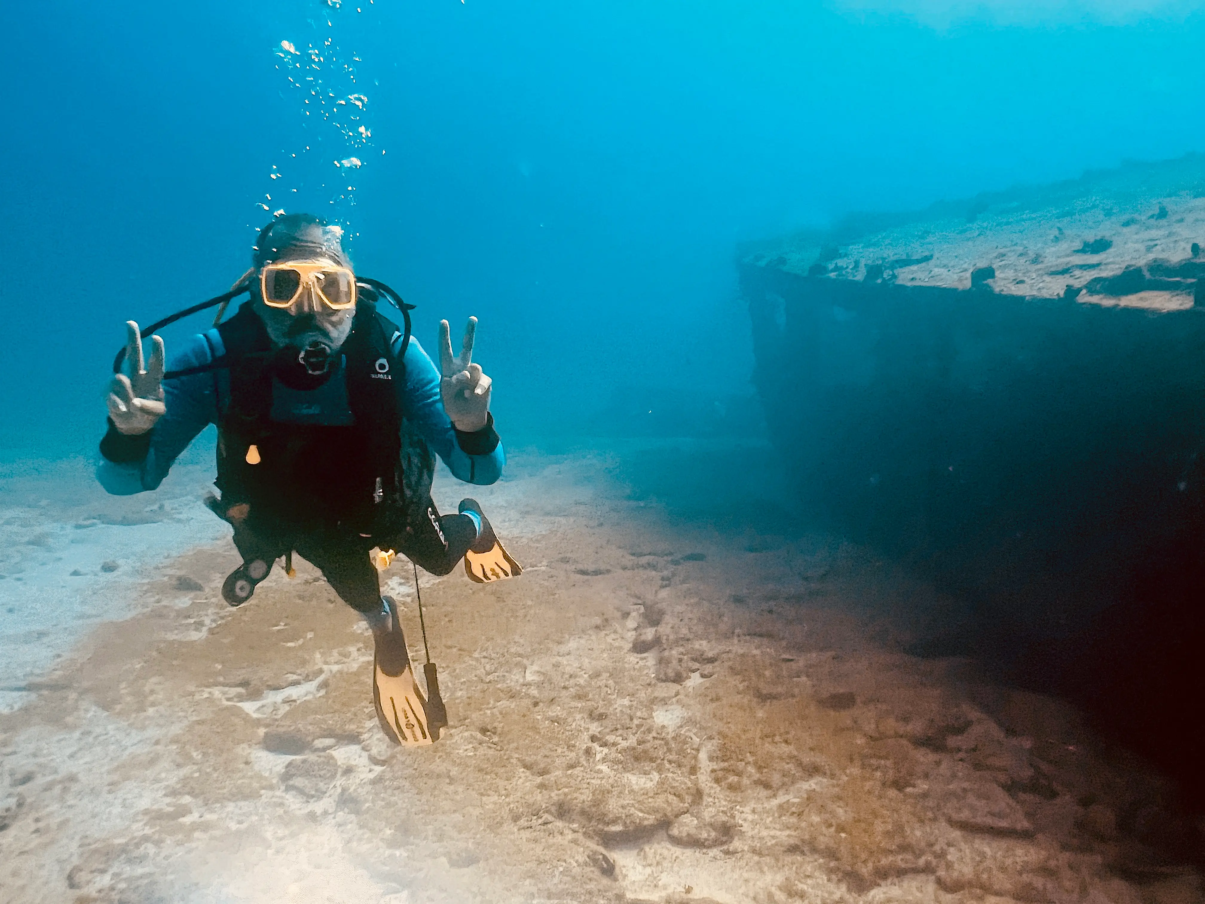 A diver makes a peace sign while exploring a wreck, set against a sandy seabed and clear water.